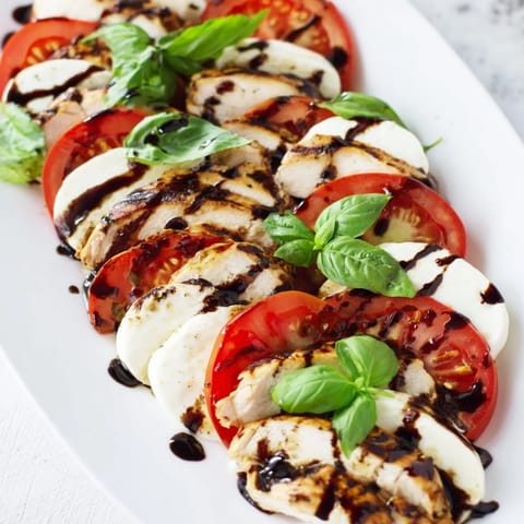 Sliced grilled chicken rests on fresh mozzarella and ripe tomato slices, garnished with basil and a glossy balsamic drizzle.
